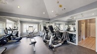 This fitness center at 1004 Meadow Creek Dr in Irving, Texas, offers a well-appointed workout facility across a single floor. The main exercise area is characterized by soaring vaulted ceilings with recessed lighting and a ceiling fan, creating an open and airy atmosphere. Neutral gray walls are complemented by large, white-trimmed windows that flood the space with natural light and provide views of the surrounding greenery. Full-length wall mirrors visually expand the room while allowing users to monitor their form during workouts. The facility is equipped with a comprehensive selection of Life Fitness cardio machines, including treadmills, ellipticals, and a stationary bike, as well as strength and resistance training equipment such as a rowing machine and weight benches. Durable rubber flooring covers the exercise area, providing both safety and comfort. A wall-mounted flat-screen television adds an entertainment element for cardio sessions. An adjacent room houses a tiled shower area, finished in classic white subway tile with a decorative diamond-pattern border accent, safety grab bars, and a wooden shower bench, offering a convenient post-workout rinse facility. French doors connect distinct zones within the space, enhancing functionality and flow throughout this compact but comprehensive fitness center.