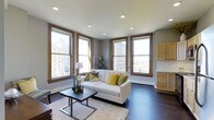 This single-floor space offers a well-designed open layout that seamlessly connects the living and kitchen areas. The living room is anchored by a clean-lined white sofa accented with green and yellow throw pillows, complemented by a minimalist rectangular coffee table resting on a textured area rug over rich, dark hardwood floors. Recessed ceiling lights provide even illumination throughout. A standout feature of the living area is its multiple large windows with warm wood trim on three sides, framing sweeping views of the surrounding urban skyline and flooding the space with abundant natural light. The neutral gray walls create a versatile backdrop that enhances the bright, airy atmosphere. Adjacent to the living area, the kitchen is partially visible and features light maple cabinetry, white subway tile backsplash, and matching light-toned finishes that contrast elegantly with the darker flooring. The overall ambiance is modern yet warm, combining urban sophistication with a welcoming, livable character well-suited to city living.