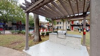 This residential community showcases a classic brick exterior complemented by warm stucco accents, black-shuttered windows, and well-maintained landscaping throughout the grounds. The multi-building layout is connected by concrete and paver walkways that wind through manicured greenery, mature trees, and neatly bordered garden beds, creating a cohesive and welcoming outdoor environment.

A standout feature of the community is the resort-style swimming pool, surrounded by a spacious brick-paver pool deck furnished with blue lounge chairs and umbrella-shaded tables for relaxed outdoor enjoyment. Adjacent to the pool, a dedicated outdoor kitchen area sits beneath a substantial wood pergola, anchored by mature trees growing through the structure. The built-in stainless steel grill and stone-clad counter provide an ideal setting for outdoor entertaining.

The courtyard areas between buildings offer additional gathering space, with paver patios, outdoor seating, and shade trees that create a pleasant communal atmosphere. Residential units feature private balconies and ground-floor patios, offering direct access to the surrounding green spaces. The architectural design balances traditional brick craftsmanship with functional outdoor amenities, resulting in a community that is both visually appealing and well-suited for outdoor living.