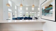 Located at 4020 Chartwell Court in Spring Hill, Tennessee, this single-floor community clubhouse offers a generously sized, open-plan layout designed for comfort and social interaction. The space is immediately distinguished by its dramatic double-height ceilings and an expansive wall of floor-to-ceiling windows with transom panels, flooding the interior with natural light and offering views of the surrounding outdoor pool and landscaping.

Underfoot, light-toned herringbone hardwood flooring runs throughout, adding a refined yet welcoming character to the space. The main gathering area features multiple seating arrangements, including a lounge zone with a tan leather sectional sofa and accent chair set against a slate-blue accent wall, as well as clusters of navy blue upholstered chairs arranged around a central rug. A dining area with round white tables and modern black chairs offers additional flexible seating.

A stylish kitchenette serves the space, complete with warm wood cabinetry, white subway tile backsplash, a quartz countertop bar with bar stool seating, pendant lighting, and open shelving. Large windows within the kitchenette frame outdoor views and bring in further natural light.

Curated artwork adorns the bright white walls throughout, and lush indoor plants punctuate the space with organic warmth. The overall ambiance is clean, modern, and inviting.