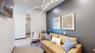 This thoughtfully designed single-floor office suite offers a compact yet highly functional layout that maximizes every square foot. The space opens into a stylish lounge area anchored by a warm caramel leather sofa, coordinating accent chairs, and a striking bold-blue feature wall adorned with framed artwork. Overhead, a sculptural linear LED chandelier adds a dramatic, modern focal point. Adjacent to the lounge, a dedicated conference room features a round wood table surrounded by cream upholstered chairs, a wall-mounted display screen, and matching linear lighting — ideal for focused meetings. The dining area offers a biophilic design touch, with a slatted wood accent wall showcasing hanging greenery panels alongside banquette seating and rounded white tables, creating a refreshing, nature-inspired atmosphere. A well-appointed kitchenette with light gray cabinetry, white quartz countertops, open shelving, and polished concrete flooring provides practical amenities throughout the workday. The open workspace area, visible in the dollhouse view, accommodates multiple workstations and benefits from natural light through perimeter windows. Connecting hallways ensure seamless flow between all areas, unifying the suite's clean, professional, and inviting character.
