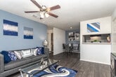 This single-floor unit presents a cohesive navy blue and grey design theme carried consistently throughout its rooms. The living area serves as the centerpiece of the space, featuring a charcoal upholstered sofa accented with an array of navy, silver, and patterned throw pillows. Matching table lamps flank the sofa, and two navy and silver abstract canvas artworks anchor the grey accent wall above. A ceiling fan with a light fixture provides overhead comfort and circulation.

The galley-style kitchen is efficiently laid out with grey cabinetry lining both walls, white countertops, and dark hardwood-style flooring. Black appliances, including a refrigerator, electric range, and dishwasher, complement the grey cabinetry, while a double stainless steel sink and a wall-mounted rack add functional detail.

The bedroom is generously sized with plush carpeting, multiple windows dressed in layered navy curtains, and a navy upholstered headboard with silver nail-head trim. Open doorways connect to a walk-in closet and additional storage areas, enhancing the room's practicality.

The bathroom features grey cabinetry with a light stone-look countertop, a wide vanity mirror with overhead lighting, an overhead medicine cabinet, and a shower-tub combination with a decorative curtain. Dark hardwood-style flooring ties the bathroom's aesthetic together.