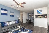 This single-floor unit presents a cohesive navy blue and grey design theme carried consistently throughout its rooms. The living area serves as the centerpiece of the space, featuring a charcoal upholstered sofa accented with an array of navy, silver, and patterned throw pillows. Matching table lamps flank the sofa, and two navy and silver abstract canvas artworks anchor the grey accent wall above. A ceiling fan with a light fixture provides overhead comfort and circulation.

The galley-style kitchen is efficiently laid out with grey cabinetry lining both walls, white countertops, and dark hardwood-style flooring. Black appliances, including a refrigerator, electric range, and dishwasher, complement the grey cabinetry, while a double stainless steel sink and a wall-mounted rack add functional detail.

The bedroom is generously sized with plush carpeting, multiple windows dressed in layered navy curtains, and a navy upholstered headboard with silver nail-head trim. Open doorways connect to a walk-in closet and additional storage areas, enhancing the room's practicality.

The bathroom features grey cabinetry with a light stone-look countertop, a wide vanity mirror with overhead lighting, an overhead medicine cabinet, and a shower-tub combination with a decorative curtain. Dark hardwood-style flooring ties the bathroom's aesthetic together.