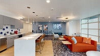This generously sized single-floor amenity space offers a thoughtfully designed layout that blends functionality with contemporary style. The open-plan common area serves as the heart of the space, anchored by a sleek kitchen featuring gray cabinetry, a bold blue-and-white hexagonal tile backsplash, stainless steel appliances, and a large white quartz island with leather barstool seating — ideal for casual gatherings or informal meetings.

Adjacent to the kitchen, a lounge area is furnished with vibrant orange sofas, accent chairs, and low wood-and-metal side tables, all set against a calming blue accent wall adorned with contemporary artwork and a wall-mounted flat-screen display. Circular pendant lighting adds a modern touch overhead, while patterned area rugs define the various seating zones across light hardwood and carpeted flooring.

A bright game and recreation area is bathed in natural light from expansive floor-to-ceiling gridded windows, offering outdoor views and housing a shuffleboard table alongside comfortable lounge seating. High-top tables and bar-height seating along the walls provide additional casual work or social spots.

A long, well-lit hallway connects the various zones, lined with artwork and leading to additional multipurpose or flex rooms. The overall ambiance is polished, welcoming, and energetic — well-suited for a residential or mixed-use community amenity floor.