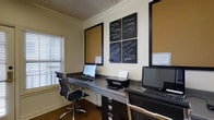 This single-floor business center offers a functional and focused environment designed to meet the everyday professional needs of its users. The room features a long, dark-finished built-in desk that runs along the wall, providing ample workspace for multiple users simultaneously. Equipped with desktop computers and a shared printer, the space is set up to handle a range of tasks from document printing to general computing. Ergonomic black office chairs offer comfortable seating at each workstation. The walls are adorned with a combination of cork bulletin boards and bold black chalkboard panels, which display community notices, helpful reminders, and welcoming messages that contribute to a friendly, communal atmosphere. Crown molding and a decorative ceiling add a touch of classic architectural character to the room. Natural light filters in through a window fitted with white horizontal blinds, keeping the space bright without glare. The warm wood-toned flooring grounds the room with a clean, professional aesthetic. Overall, the space balances practicality with charm, making it an inviting and efficient resource center.