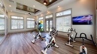 Located at 50 Cheryl Lane in Summerville, SC, this single-floor fitness center is thoughtfully designed to support a well-rounded exercise routine. The space is divided into two distinct areas connected by large glass-paned sliding doors, creating an open and airy atmosphere while allowing flexibility in how the facility is used.

The cycling studio is a standout feature, accommodating multiple stationary spin bikes arranged for group or individual sessions. A wall-mounted flat-screen TV enhances the workout experience, while a striking tray ceiling with a rich wood inlay and a ceiling fan adds both character and comfort. Large windows fitted with blinds line the walls, flooding the room with natural light and offering views of the surrounding outdoor greenery.

Adjacent to the cycling area is a functional training zone, equally well-lit with an abundance of windows and recessed lighting. This section is equipped with a Smart Fitness storage rack holding a variety of exercise balls, medicine balls, resistance tools, and yoga mats, providing everything needed for a diverse strength and flexibility workout. A second mounted flat-screen TV is positioned on the wall for added convenience.

Throughout both areas, durable hardwood-style flooring ties the space together with a clean, modern aesthetic. The overall design balances practicality with an inviting ambiance, making this fitness center a well-appointed destination for residents seeking an active lifestyle.