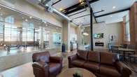 This community room showcases a sophisticated blend of industrial and contemporary design across a generously proportioned, open-plan layout. Soaring ceilings with exposed steel beams and visible ductwork lend an authentic industrial character, while warm hardwood flooring and neutral walls create an inviting atmosphere throughout.

The lounge area serves as the social centerpiece, furnished with rich brown leather sofas and armchairs arranged around a central coffee table. A linear fireplace set against a textured stone accent wall anchors the seating zone, accompanied by a wall-mounted television for entertainment. Pendant drum lighting adds a refined touch overhead.

Adjacent to the lounge, a fully equipped kitchen features white shaker-style cabinetry, granite countertops, stainless steel appliances including a refrigerator, dishwasher, and microwave, and a large island with bar seating — ideal for hosting gatherings.

A glass-walled game room partitioned by full-height sliding glass doors provides a flexible, semi-private space accommodating a billiard table and additional seating. Original exposed brick walls and oversized industrial-style windows allow abundant natural light and outdoor views, reinforcing the building's heritage character.