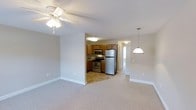 This single-floor space showcases an open-concept design that seamlessly connects the living area to the kitchen. The living area is finished with soft, neutral-toned carpeting and features a ceiling fan with integrated lighting, contributing to a comfortable and functional atmosphere. Clean white baseboards and light gray walls create a bright, cohesive aesthetic throughout.

The kitchen is accessible via an open passthrough and is outfitted with warm-toned wooden cabinetry, stainless steel appliances including a gas range, microwave, and full-size refrigerator, and tile flooring that distinguishes it from the carpeted living space. A pendant light fixture hangs above, adding a subtle decorative touch.

A hallway visible beyond the kitchen leads to additional areas of the unit, with a door at the far end allowing natural light to filter through. The overall layout is practical and efficiently designed, with a textured ceiling overhead throughout. The neutral color palette and consistent finishes give the space a clean, move-in-ready appearance suitable for everyday living.
