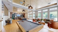 Located at 40 Pallet Rd in Candler, NC, this thoughtfully designed single-floor community amenity space offers a welcoming and versatile environment for socializing and recreation. The open-plan layout is anchored by warm light wood flooring and a striking tongue-and-groove wood ceiling, which lends a rustic yet refined character to the interior throughout.

A full-size billiard table serves as a central focal point in the recreation area, suspended beneath industrial-style pendant lighting that adds character to the space. Adjacent to this, a generously sized bar counter with a white quartz surface and bold teal cabinetry is set against a dramatic slate-toned feature wall with metallic veining, flanked by open display shelving. This gathering bar area is equipped with a beverage station and is illuminated by a cascading cluster of Edison bulb pendants.

The lounge area is furnished with plush, earth-toned seating—including a sage green sofa and cognac leather accent chairs—arranged around a live-edge wood coffee table, creating an inviting retreat bathed in natural light. Floor-to-ceiling windows and French doors on multiple walls provide sweeping outdoor views of the surrounding greenery and outdoor amenities, while beadboard wainscoting and white trim details throughout deliver a clean, coastal-inspired aesthetic. A dedicated dining area with dark wood tables and upholstered chairs rounds out the space's multifunctional appeal.