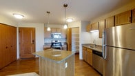 This single-floor space centers around a well-appointed kitchen designed for both functionality and comfort. The kitchen features a prominent central island topped with warm-toned granite countertops, providing generous prep space and casual seating potential. Pendant lighting hangs above the island, adding focused illumination and a touch of style.

The cabinetry throughout is finished in light honey-toned wood, offering a cohesive and warm aesthetic. Stainless steel appliances, including a range with electric cooktop, built-in microwave, dishwasher, and refrigerator, provide modern convenience. A dedicated open shelving unit offers versatile display and storage options.

The sink area is positioned along a secondary counter run, complete with additional cabinetry below and under-cabinet lighting for practical workspace visibility. Hardwood-style flooring runs throughout, contributing warmth and easy maintenance. Natural and recessed overhead lighting keeps the space bright and welcoming. An entryway is visible to the side, integrating the kitchen seamlessly into the broader single-level layout.