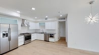 This single-floor space showcases a clean, modern aesthetic defined by a thoughtfully designed open kitchen and bright, airy interiors. The kitchen is a standout feature, outfitted with flat-front white cabinetry that extends to the ceiling, offering generous storage. Dark stone countertops provide a striking contrast against the white surfaces, while a full suite of stainless steel appliances — including a gas range with a wall-mounted hood, a dishwasher, and a built-in microwave — caters to both everyday cooking and entertaining. A frosted glass cabinet adds a subtle design detail, breaking up the clean lines of the upper cabinetry. Recessed lighting is installed throughout, ensuring consistent, well-distributed illumination across the space. Light-toned hardwood flooring runs continuously, lending warmth and cohesion to the interior. The crisp white walls and high, smooth ceilings amplify the sense of openness and space. A doorway leads to an adjacent room or corridor, suggesting a functional and well-connected floor plan. Overall, the space exudes a fresh, minimalist character well-suited for contemporary living.