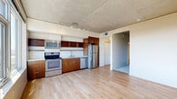 Located at 11011 NE 9th St in Bellevue, WA, this one-bedroom, one-bathroom unit offers a well-designed layout across a single floor. The open-concept living, dining, and kitchen area serves as the heart of the space, featuring light hardwood flooring, an exposed concrete ceiling, and track lighting. The kitchen is equipped with stainless steel appliances — including a refrigerator, dishwasher, range, and microwave — warm wood cabinetry, open shelving, and a clean white countertop. A large picture window in the living area floods the space with natural light and offers views of the surrounding Bellevue skyline. A well-lit hallway connects the main living area to the private bedroom, which features carpeted flooring, a sliding closet door, and an exposed concrete ceiling that adds an industrial character. The bathroom includes wood-toned floating cabinetry, a white countertop with an undermount sink, a large mirror, and light hardwood-style flooring. An in-unit laundry area is also conveniently combined within the bathroom. The overall aesthetic blends modern industrial elements with warm wood accents, creating a clean and functional living environment in the heart of Bellevue.