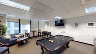 This single-floor, 2-bedroom, 2-bathroom space includes a well-appointed game and recreation room designed for entertaining and leisure. The game room serves as a central gathering area, anchored by a full-sized pool table with a dark felt surface and complete with cues and racked balls, ready for play. Adjacent to the pool table sits a foosball table, adding another layer of recreational options for guests and residents alike.

A wall-mounted flat-screen television is positioned prominently, making the space suitable for both gaming and viewing. The room also features a tall bistro-style table with matching chairs tucked near the windows, offering a casual spot for conversation or refreshments. A small seating area with a round coffee table and upholstered chairs provides additional lounge space.

The room is finished with neutral-toned carpeting and white walls adorned with framed artwork, contributing to a clean, functional aesthetic. A drop-panel ceiling with recessed fluorescent lighting ensures even illumination throughout. Windows along one wall allow natural light to filter in, enhancing the overall atmosphere of the space.