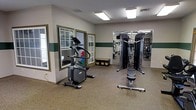 This fitness room offers a practical and well-organized space designed to support a range of exercise routines. The room features durable rubber flooring suitable for workout activity, and is finished with neutral walls accented by a dark green horizontal stripe, lending a clean and classic aesthetic throughout.

Natural light enters through a set of white-framed windows and a glass-paned door, brightening the space alongside overhead fluorescent lighting that ensures consistent illumination. A large wall mirror on one side of the room visually expands the space while allowing users to monitor their form during exercise.

The room is equipped with a standing elliptical cardio machine and a multi-function cable strength training station, providing options for both cardiovascular and resistance-based workouts. Additional storage is available via a multi-compartment organizer near the entry door, keeping the space tidy and functional.

Overall, the fitness room presents a straightforward and accessible environment, suitable for residents or occupants looking to maintain an active lifestyle without leaving the property.