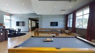 This contemporary single-floor amenity space is thoughtfully designed for recreation and social interaction. The game room serves as the centerpiece, featuring a full-size pool table in the foreground and a ping-pong table positioned nearby, making it an ideal setting for friendly competition. A wall-mounted flat-screen television is prominently displayed against a textured grey accent wall, adding an entertainment focal point to the room. The space is finished with recessed ceiling lighting that provides a clean, modern ambiance, complemented by a tray ceiling detail that adds architectural depth. Natural light filters in through tall windows dressed with dark curtains, offering a pleasant connection to the outdoors. The flooring is a smooth, light-toned surface that ties the room together cohesively. Neutral-toned seating is tucked along the perimeter, providing comfortable areas to relax and socialize. A partially visible adjacent room with glass partitions hints at additional functional spaces beyond. The overall design palette of greys, browns, and warm neutrals creates a sophisticated yet inviting atmosphere well-suited for communal use.