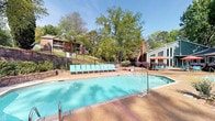 This outdoor amenity space centers around a generously sized swimming pool with crystal-clear blue water, accessible via stainless steel entry rails on both ends. The pool deck is finished in a textured aggregate surface accented by brick-inlaid borders, providing both visual appeal and safe footing. Depth markers are clearly labeled along the pool's edge, reflecting attention to safety standards throughout the area.

A row of teal-colored lounge chairs lines one side of the deck, offering ample space for sunbathing and relaxation. Additional seating is scattered throughout the area, including coordinating teal chairs positioned along raised patio terraces. A shaded dining area with umbrella tables is visible near an adjacent structure, expanding the outdoor entertainment options.

The surrounding landscape is thoughtfully designed, featuring tiered retaining walls constructed from large, textured masonry blocks. These walls create defined planting beds filled with mulch, ornamental shrubs, and flowering plants, adding layers of greenery and color. Wrought iron fencing tops the upper retaining walls, providing both security and a refined architectural detail.

Towering mature trees frame the entire space, casting dappled shade and creating a private, park-like atmosphere. The lush canopy, combined with the well-maintained grounds and brick-accented hardscaping, contributes to a serene and inviting outdoor environment suited for both active recreation and leisurely relaxation.