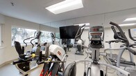 The fitness center is a bright, single-level space designed to support a well-rounded cardio workout routine. The room is lined with floor-to-ceiling mirrors along one wall, which visually expand the space and allow users to monitor their form during exercise. A large flat-screen television is mounted centrally on the mirrored wall, providing entertainment during workouts.

Natural light fills the room through windows that overlook the street outside, contributing to an open and energizing atmosphere. Overhead, recessed panel lighting ensures consistent illumination throughout the space during all hours.

The fitness center is outfitted with a variety of cardio equipment, including elliptical trainers, stationary bikes, and stair-climbing machines, each equipped with individual digital display consoles for tracking workout metrics. The equipment is arranged to allow comfortable movement between machines. The flooring is light-toned and easy to maintain, complementing the clean, neutral color palette of the walls and ceiling. Overall, the space offers a functional and comfortable environment for cardiovascular training.