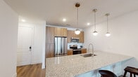 This contemporary single-floor residence is designed with a clean, modern aesthetic and thoughtful attention to detail throughout. The open-concept kitchen serves as the centerpiece of the space, anchored by a sweeping granite-topped island with seating for multiple bar stools. Warm wood cabinetry extends to the ceiling, complemented by a full suite of stainless steel appliances including a French door refrigerator, electric range, and built-in microwave. A series of globe pendant lights hang overhead, adding a stylish finishing touch. Wood-look flooring flows through the main living areas, creating a cohesive and inviting atmosphere. The living area features multiple windows and a private exterior door that allow ample natural light to fill the room, contributing to an airy, open feel. The bedroom is comfortably sized with large windows, carpeted flooring, and a ceiling fan for added comfort. The primary bathroom is well-appointed with a granite-topped wood vanity, a glass-enclosed shower, and direct access to a dedicated walk-in closet, providing generous storage. Throughout the residence, neutral tones, recessed lighting, and consistent finishes create a polished and cohesive living environment.