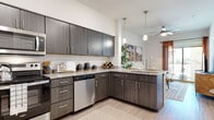 This single-floor residence showcases a thoughtfully designed layout that balances modern aesthetics with everyday functionality. The kitchen serves as a central highlight, featuring dark wood-grain cabinetry extending to elevated heights, granite-look countertops, and a classic white subway tile backsplash. A full suite of stainless steel appliances — including a refrigerator, electric range, over-the-range microwave, and dishwasher — provides a complete and polished cooking environment. An L-shaped peninsula with pendant lighting above creates a natural transition into the living area, which is topped by a ceiling fan and receives ample natural light. Light wood-look flooring runs throughout the main living spaces, contributing to a clean, cohesive feel.

The bedroom offers a comfortable, carpeted retreat, highlighted by a striking landscape mural that serves as a dramatic accent wall. A large window dressed with blinds and sheer curtains allows natural light to filter softly into the room.

The bathroom maintains the design consistency found throughout the space, featuring matching dark cabinetry with granite countertops, wood-look flooring, and a tub-shower combination. The overall ambiance of the residence is contemporary and inviting, with warm neutral tones tying each space together seamlessly.