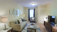 This single-floor residence offers a comfortable and well-appointed living environment designed with both style and practicality in mind. The living area serves as the heart of the space, featuring warm, soft yellow-green walls that create a calm and inviting atmosphere. Light hardwood-style flooring runs throughout, adding warmth and continuity to the space.

The living room is thoughtfully arranged with a cushioned sofa adorned with decorative pillows, complemented by a coordinating accent chair. A rectangular coffee table anchors the seating area, which is defined by a patterned area rug. Framed artwork decorates the walls, adding visual interest alongside a soft floor lamp that provides warm ambient lighting.

Natural light flows into the room through a large window dressed with dark grommet-top curtains, offering a pleasing contrast to the light-toned walls. A louvered closet door is visible, hinting at practical storage solutions integrated seamlessly into the design. The overall ambiance is serene and harmonious, with a neutral color palette that makes the space feel open, airy, and easy to personalize. The layout is efficient and cohesive, making excellent use of the available footprint while maintaining a comfortable sense of scale.