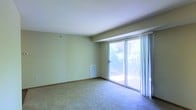 This single-floor space features a spacious main living area finished with wall-to-wall carpeting and smooth, light-toned walls that create a clean, neutral backdrop. A textured ceiling adds subtle architectural detail overhead. The standout feature of the room is a large sliding glass door framed by sheer white curtains, which floods the interior with abundant natural light and provides direct access to a private balcony. The balcony overlooks a verdant outdoor setting, with tree foliage visible beyond the railing, lending the space a pleasant connection to the outdoors. A floor-level HVAC vent is integrated into the wall, indicating a built-in climate control system. The open layout of the living area offers flexibility for a variety of furniture arrangements. The overall ambiance is bright and airy, with natural light serving as a defining characteristic of the space. The minimalist, unfurnished presentation allows the architectural elements — including the generous proportions of the main room and the prominent glazed access point to the balcony — to remain the focal points of the interior.