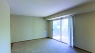 This single-floor space features a spacious main living area finished with wall-to-wall carpeting and smooth, light-toned walls that create a clean, neutral backdrop. A textured ceiling adds subtle architectural detail overhead. The standout feature of the room is a large sliding glass door framed by sheer white curtains, which floods the interior with abundant natural light and provides direct access to a private balcony. The balcony overlooks a verdant outdoor setting, with tree foliage visible beyond the railing, lending the space a pleasant connection to the outdoors. A floor-level HVAC vent is integrated into the wall, indicating a built-in climate control system. The open layout of the living area offers flexibility for a variety of furniture arrangements. The overall ambiance is bright and airy, with natural light serving as a defining characteristic of the space. The minimalist, unfurnished presentation allows the architectural elements — including the generous proportions of the main room and the prominent glazed access point to the balcony — to remain the focal points of the interior.