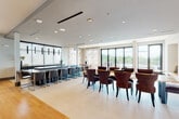 This thoughtfully designed amenity space presents a seamless blend of interior sophistication and outdoor functionality across a single level. The interior is anchored by a generous open-plan area that transitions fluidly between a formal dining zone and a relaxed lounge. Large-format light-toned tile flooring runs throughout the main gathering space, while warm hardwood flooring defines the lounge area. Floor-to-ceiling windows and glass doors flood the interior with natural light and provide direct access to an adjacent outdoor terrace, creating a strong indoor-outdoor connection.

The dining area features multiple substantial wood tables paired with upholstered seating in rich brown tones, accommodating both intimate and large-group gatherings. A sleek wet bar with a striking veined dark granite countertop and matte black cabinetry serves as a focal point, illuminated by a series of slim pendant lights. The lounge area is anchored by a textured dark accent wall housing a large-format television, complemented by hardwood shelving and warm seating.

The rooftop terrace extends the social space outdoors, finished with teak-style deck tiles and furnished with contemporary modular seating and globe pendants strung overhead, offering open views of the surrounding landscape.