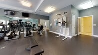 This fitness center is a thoughtfully designed, single-level space built to support a comprehensive workout experience. The room features walls painted in cool gray and soft teal tones, complemented by durable rubber flooring that provides both safety and comfort underfoot. Flush-mounted ceiling light panels illuminate the space evenly, while windows along one wall allow natural light to filter in and offer views of an outdoor pool area.

The cardio zone is anchored by a row of treadmills and elliptical machines, including SportsArt-brand equipment, positioned in front of large mirrored walls that enhance the sense of openness and allow users to monitor their form. Multiple wall-mounted flat-screen TVs are positioned for easy viewing from the cardio equipment.

The strength-training area includes a multi-station cable machine, a weight bench, a vertical knee raise station, and a dumbbell rack with free weights. Workout reference charts are posted on the wall for user guidance. A fitness center rules sign is prominently displayed near the entrance, indicating this is a shared community facility. The overall ambiance is clean, functional, and well-maintained.