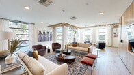 Located at 5727 North 7th Street in Phoenix, AZ, this two-story clubroom is a thoughtfully designed communal space that blends modern aesthetics with functional versatility. The ground floor makes a striking first impression with a gallery-style hallway adorned with bold, gold-framed artwork set against richly patterned blue wallcovering and warm hardwood flooring. A glass-paneled entry labeled "Clubroom" leads into a spacious, light-filled lounge featuring multiple seating arrangements, including plush sofas, rounded accent chairs, and layered area rugs. Oversized windows dressed with sheer white drapery flood the room with natural light, while a distinctive slatted wood ceiling feature adds architectural depth and visual warmth. The upper floor offers a more relaxed, entertainment-focused atmosphere, housing a full-size pool table alongside a sleek dining area with a sculptural pendant light. A second lounge area on this level features a generous sectional sofa in warm tan leather, complemented by large windows overlooking the surrounding neighborhood. An open-railing staircase connects both levels, maintaining a sense of openness throughout. The overall ambiance is contemporary and curated, with a cohesive palette of warm neutrals, natural wood tones, and carefully selected art that gives the space a polished, welcoming character.