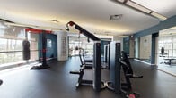 This fitness center is a comprehensively outfitted facility designed to support a full range of workout routines. The space is divided into distinct functional zones, each tailored to a specific type of training. The cardio area features a row of treadmills and stationary bikes aligned along large, floor-to-ceiling windows that invite abundant natural light and offer open views of the surrounding landscaped grounds and neighboring buildings. The strength training zone is equipped with multiple cable machines, selectorized weight stations, and free-weight accessories, providing users with a versatile array of resistance training options. A dedicated functional training area includes a boxing bag, battle ropes, medicine balls, resistance bands, and a power tower, catering to dynamic and high-intensity workouts. A cylindrical structural column wrapped in motivational typography serves as a distinctive design element and visual anchor in the space. The walls are painted in a deep teal hue, complemented by large mirrors that enhance the sense of space. Rubber flooring throughout ensures safety and durability. An adjacent lounge or entry area with built-in cabinetry, a beverage cooler, and framed artwork provides a transitional space between the gym and the rest of the facility.