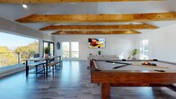 This inviting single-floor recreational space combines rustic charm with modern comfort through a thoughtfully designed open layout. The standout architectural feature is the vaulted ceiling adorned with prominent natural wood beams, which add warmth and visual height to the entire space. White shiplap walls complement the beams beautifully, creating a clean, coastal-inspired aesthetic throughout.

The main gathering area is generously sized, anchored by a solid wood pool table at its center, while a shuffleboard table provides additional entertainment along one side. A comfortable lounge area features a plush sofa, a woven jute rug, and a wall-mounted flat-screen TV, making it ideal for relaxing between games. Long communal bench tables near the windows offer additional seating for larger groups.

Natural light pours in abundantly through multiple large windows and glass doors, creating a bright and airy atmosphere. Sliding glass doors and French doors provide direct access to the outdoors, further enhancing the open feel of the space.

A well-appointed kitchenette supports the entertaining function of the room, featuring white shaker cabinetry with glass-front upper cabinets, a classic subway tile backsplash, dark countertops, an undermount sink, a beverage refrigerator, and small appliances. The durable wood-look flooring in cool gray tones ties the entire space together with a relaxed, beach-house sensibility.