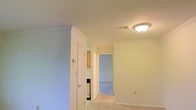 This single-floor residence offers a clean, functional layout designed for comfortable everyday living. The interior is finished with neutral, light-toned walls throughout, creating a bright and airy ambiance that makes the space feel open and welcoming. Soft carpeted flooring runs through the main living areas and into the adjoining rooms, adding warmth underfoot.

The hallway serves as a central connector, linking the living area to the kitchen and additional rooms beyond. A doorway reveals a glimpse into the kitchen, where light wood cabinetry is visible, suggesting a practical and well-organized culinary space. A thermostat mounted on the wall indicates climate control convenience for year-round comfort.

A flush-mount ceiling light fixture provides consistent overhead illumination, contributing to the overall brightness of the interior. White trim and door frames provide clean contrast against the soft wall tones, lending a tidy, well-maintained appearance throughout. The open doorway leading to a carpeted room beyond hints at a thoughtfully connected floor plan that balances privacy with accessibility. Overall, this single-level space presents a move-in ready environment with a straightforward, practical design suited for a variety of living arrangements.