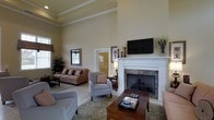 This single-story space centers around a generously proportioned living area defined by its dramatically high ceilings and clean architectural lines. Crown molding frames the upper walls, adding a refined touch to the otherwise neutral, warm-toned palette. The focal point of the room is a classic white-trimmed fireplace with a stone surround, above which a wall-mounted flat-screen television is positioned for easy viewing. The living area is furnished with a comfortable sofa and coordinating armchairs arranged around a wooden coffee table atop a patterned area rug, creating a welcoming and well-organized seating arrangement. Light tile flooring extends throughout the space, contributing to the bright and airy atmosphere. Natural light filters in through windows dressed with fabric valances, adding warmth to the interior. Decorative elements, including candlesticks, framed artwork, and potted greenery, are thoughtfully placed throughout. A doorway visible in the background hints at additional connected spaces, suggesting a functional and cohesive floor plan overall.