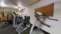 This fitness room offers a focused and functional workout environment, providing users with a dedicated space for cardiovascular exercise. The room is equipped with a selection of cardio machines, including a treadmill, an elliptical trainer, and a recumbent stationary bike, catering to a range of fitness preferences and intensity levels. A ceiling fan is mounted overhead, helping to maintain airflow and comfort during workouts. The walls are lined with mirrors, which enhance the sense of space and allow users to monitor their form while exercising. A water dispenser is conveniently positioned within the room, ensuring hydration is readily accessible. The flooring features a dark, low-pile surface well-suited for a fitness setting. Horizontal blinds on the window provide adjustable light control. Wood trim detailing along the upper walls adds a subtle finishing touch to the otherwise utilitarian space. The room's straightforward layout ensures all equipment is easily accessible and efficiently arranged, making it a practical resource for those seeking a convenient on-site fitness option.