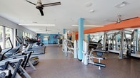 This fitness center offers a spacious, single-floor layout designed to accommodate a wide range of workout routines. The interior is defined by bold design choices, including teal structural columns and an orange accent beam that run through the ceiling, creating a modern, energetic ambiance. Walls painted in a deep teal complement the lively color scheme throughout.

The flooring consists of durable rubber gym matting, providing both safety and comfort during exercise. Ceiling fans are mounted overhead to maintain airflow throughout the space, supplemented by recessed lighting that ensures the area remains bright and well-lit.

Equipment is organized into distinct zones. A row of treadmills and cardio machines lines one side of the room, accompanied by a mounted flat-screen television. The central area houses a multi-station strength training machine, while a dedicated free weights section features a full rack of dumbbells alongside weight benches and mirrors that span the wall, allowing users to monitor their form. The overall layout promotes ease of movement between stations, making the fitness center functional and accessible for a variety of fitness levels.