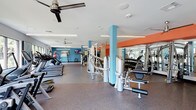 This fitness center offers a spacious, single-floor layout designed to accommodate a wide range of workout routines. The interior is defined by bold design choices, including teal structural columns and an orange accent beam that run through the ceiling, creating a modern, energetic ambiance. Walls painted in a deep teal complement the lively color scheme throughout.

The flooring consists of durable rubber gym matting, providing both safety and comfort during exercise. Ceiling fans are mounted overhead to maintain airflow throughout the space, supplemented by recessed lighting that ensures the area remains bright and well-lit.

Equipment is organized into distinct zones. A row of treadmills and cardio machines lines one side of the room, accompanied by a mounted flat-screen television. The central area houses a multi-station strength training machine, while a dedicated free weights section features a full rack of dumbbells alongside weight benches and mirrors that span the wall, allowing users to monitor their form. The overall layout promotes ease of movement between stations, making the fitness center functional and accessible for a variety of fitness levels.