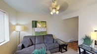 This single-floor studio is thoughtfully laid out to maximize functionality within a compact footprint. The living area serves as the central space, featuring carpet flooring, a ceiling fan with integrated lighting, and a wall-mounted air conditioning unit. A window fitted with horizontal blinds allows natural light to filter in, creating a bright and comfortable atmosphere. The space accommodates a sectional sofa, glass-top coffee table, and end table, demonstrating its practical use of available room.

The kitchen is efficiently designed with warm honey-toned wood cabinetry spanning both upper and lower sections, providing ample storage. Dark countertops contrast the cabinetry, while a white electric coil-burner range, stainless steel sink, microwave, and black refrigerator complete the appliance package. The kitchen features hardwood-style flooring, distinguishing it from the carpeted living area.

The bathroom includes a full tub and shower combination with chrome fixtures and a curtain rod, finished in clean white surroundings. The overall layout, visible in the 3D overview, reveals a straightforward open flow between the living, kitchen, and bathroom areas, making this studio a practical and self-contained living space.
