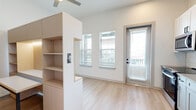 Located at 1148 North Main Street in Jacksonville, FL, this well-designed one-bedroom, one-bathroom unit offers a thoughtfully organized single-floor layout. The combined kitchen and living area serves as the heart of the space, featuring light hardwood-style flooring, large windows, and a glass-panel entry door that fills the room with natural light. A built-in shelving unit with light wood cabinetry provides both storage and a natural partition between the kitchen and living zones. The kitchen is outfitted with white shaker-style cabinetry, a white subway tile backsplash, gray quartz-style countertops, and a full suite of stainless steel appliances including a range, microwave, dishwasher, and refrigerator, all accented with matte black hardware. A hallway connects the main living area to the private bedroom, which features plush wall-to-wall carpeting and a ceiling fan. A sliding barn door with black hardware adds a contemporary design touch to the hallway. The dedicated closet offers built-in rod and shelf storage. The bathroom is finished with light wood-tone tile flooring, a gray vanity with white countertop, and a tub/shower combination clad in classic white subway tile, complemented by matte black fixtures throughout. An in-unit laundry area completes this efficient and modern residence.