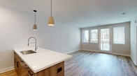 This well-appointed single-floor residence offers a thoughtfully designed open-concept layout that seamlessly connects the kitchen and living areas. Light hardwood-style flooring flows throughout the main living spaces, enhancing the bright and airy atmosphere created by large windows and a glass-panel exterior door that invites abundant natural light.

The modern kitchen serves as the heart of the home, anchored by a center island with a white quartz countertop, an undermount sink, and warm wood-toned cabinetry. Stainless steel appliances — including a gas range, over-the-range microwave, and French door refrigerator — provide a sleek, functional workspace. Pendant lighting above the island and recessed ceiling lights add warmth and dimension to the space. White upper cabinetry and a light-toned backsplash complete the clean, contemporary aesthetic.

The living area is spacious and open, benefiting from direct access to the outdoors through a glass-paneled door flanked by windows with adjustable blinds. The bedroom features plush carpeting, a generous window for natural light, and a ceiling-mounted light fixture.

The bathroom is finished with quartz countertops, a wood-toned vanity, an illuminated frameless mirror, and a glass-enclosed walk-in shower with white subway tile — delivering a clean, spa-like feel throughout.
