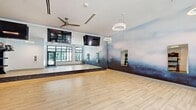 This contemporary fitness studio offers a well-appointed, open-plan exercise space designed to accommodate a wide range of workout activities. The room is finished with light-toned hardwood-style flooring that runs throughout, providing a clean and durable surface ideal for movement-based exercise. A full-length mirrored wall along one side amplifies the sense of space and serves as a functional training aid, while a dark granite-trimmed base adds a polished architectural detail.

The studio is illuminated by a combination of natural light streaming through large, black-framed floor-to-ceiling windows and stylish circular ring pendant lights suspended from the ceiling. Multiple ceiling fans contribute to air circulation and comfort during intense workouts. Several wall-mounted flat-screen TVs are positioned for easy viewing from anywhere in the room.

One wall features a striking artistic mural with a soft, atmospheric sky motif in cool blue and grey tones, adding a distinctive visual element to the space. A dedicated equipment storage area along the wall houses a multi-tiered rack stocked with TRX suspension trainers, stability balls, medicine balls, dumbbells, and resistance bands, ensuring all essential training tools are readily accessible. The studio's entry features a glass-panel door with direct views to an adjacent outdoor area, reinforcing a bright and energizing atmosphere throughout.