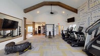 This single-floor fitness center in Lewis Center, Ohio offers a comprehensive workout environment designed with both functionality and style in mind. The main exercise area is defined by soaring vaulted ceilings adorned with rustic wood beam accents and a ceiling fan, creating an open, airy atmosphere that feels spacious and inviting. Patterned carpet tiles in gray and yellow cover the main floor, adding a modern aesthetic throughout the space.

The cardio zone is well-appointed with a lineup of TRUE brand treadmills, ellipticals, and recumbent bikes, each equipped with individual screens for an engaging workout experience. Multiple wall-mounted flat-screen televisions are positioned throughout for additional entertainment. A large arched window and a bank of full-length windows flood the space with natural light and provide views of an adjacent outdoor pool area.

A motivational typography accent wall energizes the cardio section, while a large framed mirror on the opposite wall enhances the sense of space. The strength training area transitions to hardwood flooring and features a power rack, adjustable benches, a full rack of dumbbells, cable machines, a functional trainer, and a tire sled for functional fitness. Decorative wood columns help define the zones while maintaining an open floor plan throughout.