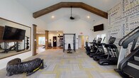This single-floor fitness center in Lewis Center, Ohio offers a comprehensive workout environment designed with both functionality and style in mind. The main exercise area is defined by soaring vaulted ceilings adorned with rustic wood beam accents and a ceiling fan, creating an open, airy atmosphere that feels spacious and inviting. Patterned carpet tiles in gray and yellow cover the main floor, adding a modern aesthetic throughout the space.

The cardio zone is well-appointed with a lineup of TRUE brand treadmills, ellipticals, and recumbent bikes, each equipped with individual screens for an engaging workout experience. Multiple wall-mounted flat-screen televisions are positioned throughout for additional entertainment. A large arched window and a bank of full-length windows flood the space with natural light and provide views of an adjacent outdoor pool area.

A motivational typography accent wall energizes the cardio section, while a large framed mirror on the opposite wall enhances the sense of space. The strength training area transitions to hardwood flooring and features a power rack, adjustable benches, a full rack of dumbbells, cable machines, a functional trainer, and a tire sled for functional fitness. Decorative wood columns help define the zones while maintaining an open floor plan throughout.