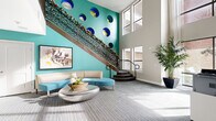 Imperial Lofts presents a thoughtfully designed communal space that makes an immediate statement upon entry. The front entrance is defined by full-height glass doors framed in dark metal, welcoming residents and visitors into a bright, open corridor with smooth flooring, sleek wall-mounted artwork, and elegant bench seating along the hallway. Lush indoor trees and potted plants add a natural warmth throughout.

The centerpiece of the space is a dramatic double-height lobby featuring a striking teal accent wall adorned with a series of bold circular sculptural elements in blue and gold. A decorative staircase with an intricate laser-cut metal railing rises along the wall, further enhancing the artistic character of the room. Curved, contemporary seating in coordinating teal tones surrounds a sculptural metallic coffee table, while a large framed artwork grounds the lounge area.

The adjacent leasing office is equally impressive, bathed in natural light from oversized multi-pane windows that frame views of the exterior greenery. Workstations with lime-green privacy panels and ergonomic seating provide a functional yet stylish workspace. The consistent use of patterned carpet, neutral walls, and vibrant accent colors ties the entire space together into a cohesive, modern aesthetic.
