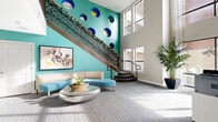 Imperial Lofts presents a thoughtfully designed communal space that makes an immediate statement upon entry. The front entrance is defined by full-height glass doors framed in dark metal, welcoming residents and visitors into a bright, open corridor with smooth flooring, sleek wall-mounted artwork, and elegant bench seating along the hallway. Lush indoor trees and potted plants add a natural warmth throughout.

The centerpiece of the space is a dramatic double-height lobby featuring a striking teal accent wall adorned with a series of bold circular sculptural elements in blue and gold. A decorative staircase with an intricate laser-cut metal railing rises along the wall, further enhancing the artistic character of the room. Curved, contemporary seating in coordinating teal tones surrounds a sculptural metallic coffee table, while a large framed artwork grounds the lounge area.

The adjacent leasing office is equally impressive, bathed in natural light from oversized multi-pane windows that frame views of the exterior greenery. Workstations with lime-green privacy panels and ergonomic seating provide a functional yet stylish workspace. The consistent use of patterned carpet, neutral walls, and vibrant accent colors ties the entire space together into a cohesive, modern aesthetic.