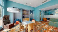Located at 3301 Glenn McConnell Pkwy in Charleston, SC, this single-floor community clubhouse and leasing space makes an immediate impression with its bold, contemporary design. Vivid teal walls set an energetic tone throughout, complemented by a striking multicolored carpet featuring a dynamic abstract pattern in orange, teal, and cream tones that anchors the entire space.

The main lounge area features a generously sized sectional sofa upholstered in a teal and gold leaf-print fabric, paired with accent chairs in orange and mustard tones. A round marble-topped coffee table and modern pendant lighting add refined touches to the lively palette. Floor-to-ceiling windows and arched transoms flood the space with natural light while offering direct views of an outdoor pool, seamlessly connecting the interior to the exterior amenity area.

A dedicated workstation area includes computer desks and task chairs, providing a functional workspace within the communal setting. Along one wall, a built-in refreshment nook features a coffee station, mini-refrigerator, and cabinetry framed by pendant lights, flanked on either side by cozy upholstered alcove seating with teal cushions. Glass-partitioned offices or conference rooms offer semi-private spaces for meetings or leasing activities. Recessed lighting throughout ensures a well-lit, welcoming atmosphere at any time of day.