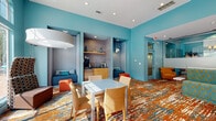 Located at 3301 Glenn McConnell Pkwy in Charleston, SC, this single-floor community clubhouse and leasing space makes an immediate impression with its bold, contemporary design. Vivid teal walls set an energetic tone throughout, complemented by a striking multicolored carpet featuring a dynamic abstract pattern in orange, teal, and cream tones that anchors the entire space.

The main lounge area features a generously sized sectional sofa upholstered in a teal and gold leaf-print fabric, paired with accent chairs in orange and mustard tones. A round marble-topped coffee table and modern pendant lighting add refined touches to the lively palette. Floor-to-ceiling windows and arched transoms flood the space with natural light while offering direct views of an outdoor pool, seamlessly connecting the interior to the exterior amenity area.

A dedicated workstation area includes computer desks and task chairs, providing a functional workspace within the communal setting. Along one wall, a built-in refreshment nook features a coffee station, mini-refrigerator, and cabinetry framed by pendant lights, flanked on either side by cozy upholstered alcove seating with teal cushions. Glass-partitioned offices or conference rooms offer semi-private spaces for meetings or leasing activities. Recessed lighting throughout ensures a well-lit, welcoming atmosphere at any time of day.