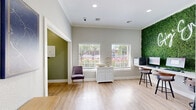 Located at 1800 Holleman Dr in College Station, TX, this single-floor office space delivers a polished and functional environment designed for productivity and collaboration. The layout is thoughtfully organized, featuring an open main area anchored by a striking floor-to-ceiling living green wall adorned with a "Gig 'Em" neon sign — an eye-catching focal point that adds energy and character to the workspace. Beneath it, a row of desktop computer workstations with bar-height seating provides a dedicated area for focused work or resident use.

Light hardwood flooring runs throughout, complementing the soft gray and sage-toned walls and lending the space a clean, modern aesthetic. Large windows along the exterior wall invite an abundance of natural light, framing views of the lush landscaping outside. Crown molding and recessed lighting add refined architectural detail overhead.

A private meeting alcove off the main area features a round white table, upholstered seating, and a wall-mounted whiteboard — ideal for small group discussions or one-on-one consultations. A console table with framed map art and a welcoming entry door complete the layout, creating a space that balances professional utility with an inviting, community-oriented atmosphere.