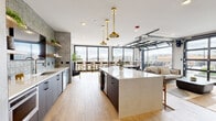 Located at 1170 Temple Dr in Sugar Hill, Georgia, this contemporary amenity space is designed to impress with a seamless blend of modern aesthetics and functional design. The open-concept kitchen serves as a centerpiece, showcasing dark cabinetry contrasted by a large quartz waterfall island, a textured tile backsplash, stainless steel appliances, and a series of elegant brass pendant lights that add a warm, sophisticated glow. Wood-look tile flooring runs throughout, unifying the space with a clean, cohesive finish. Floor-to-ceiling black-framed windows flood the interior with natural light and frame expansive views of the surrounding Sugar Hill skyline. A standout architectural feature is the retractable glass garage doors that open the lounge area directly to the outdoor terrace, creating a fluid indoor-outdoor living experience. The lounge is furnished with neutral sofas, wicker accent chairs, and sculptural coffee tables, offering a relaxed yet refined atmosphere. The expansive outdoor terrace is thoughtfully appointed with bistro-style seating, high bar tables, lush planters, and strung market lights, making it an ideal setting for both casual gatherings and social events. A separate glass-walled room provides a more private setting for meetings or focused work.