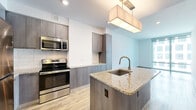 Located on Tysons Central Street in Vienna, VA, this contemporary 2-bedroom, 2-bathroom residence offers a well-designed single-floor layout with modern finishes throughout. The open-concept kitchen and living area serves as the heart of the home, anchored by a granite-topped island with an undermount sink and stylish pendant lighting. The kitchen is outfitted with sleek, gray wood-grain cabinetry, a subway tile backsplash, and a full suite of stainless steel appliances, including a refrigerator, range, and built-in microwave. Light wood-tone flooring runs throughout the main living areas, enhancing the bright and airy atmosphere. Floor-to-ceiling windows in the living room invite abundant natural light and provide views of the surrounding urban landscape. The spacious primary bedroom features plush carpeting and generous proportions, accompanied by a walk-in closet with built-in wire shelving for ample storage. The second bedroom is equally comfortable with soft carpeting and clean white walls. Both bathrooms are finished with granite-topped vanities and gray cabinetry, with one bathroom featuring a tiled tub-shower combination and brushed nickel fixtures. The overall design is clean, modern, and cohesive, making this a well-appointed urban residence.
