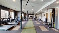 Located at 7325 Quivira Rd in Shawnee, Kansas, this professionally designed fitness center offers a comprehensive workout facility across a single, generously proportioned floor. The space is thoughtfully organized into distinct training zones, each catering to different exercise disciplines.

The main workout area features a striking combination of patterned carpet tiles in neutral tones with a central green turf runner, providing functional flooring for a variety of training activities. Structural columns are integrated throughout the open layout, complementing the modern industrial aesthetic. Ceiling fans ensure comfortable airflow across the expansive floor plan.

The cardio zone is lined with a robust selection of treadmills and ellipticals positioned along windowed walls, allowing natural light to fill the space. The strength training area houses a full dumbbell rack, a rowing machine, resistance equipment, and a power rack with accessories including medicine balls and a trampoline.

A glass-enclosed studio room with warm hardwood flooring, a richly colored ceiling, dual wall-mounted televisions, and built-in shelving provides an ideal space for group classes, stretching, or yoga. Large mirrors and a glass wall connect this room visually to the main floor.

The overall ambiance is modern and motivating, with dark accent walls, recessed lighting, and a well-curated equipment layout that supports a full spectrum of fitness training needs.